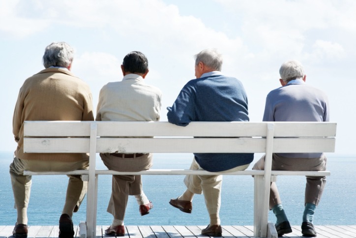 A group of four senior men sitting on a bench listing to one another complain.