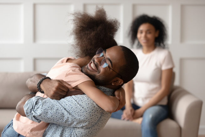 Happy dad embracing daughter cuddling at home, smiling black father hugging cute little kid girl bonding enjoy time together, daddy child reunion, fatherhood, shared child custody concept