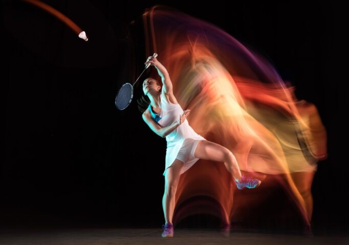 Young woman playing badminton isolated on black background in mixed light. Female model with the racket in action, motion in game with the fire shadows. Concept of sport, movement, healthy lifestyle.