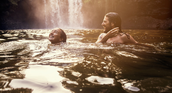 Two young adults happily playing in a river, one person up to their neck, the other just about their chest. Waterfall in the background pours into the river.