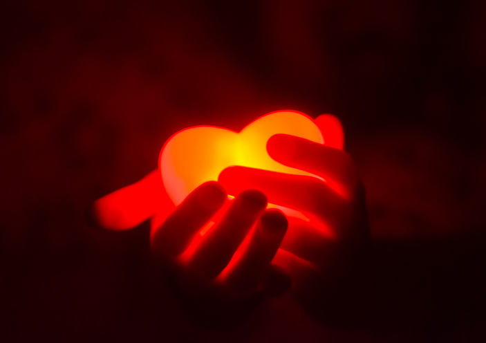 Hands gently holding red, glowing, heart-shaped light in the dark.