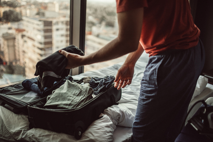 Photo of a young man, packing his wardrobe on bed by the window.