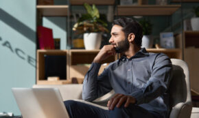 Young thoughtful divorced man looking away relaxing sitting on chair in modern office lobby with laptop, dreaming of success, planning.