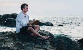 Man sits on coastal rocks and writes in a journal as the waves crash.
