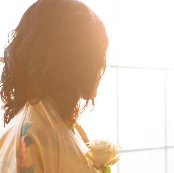 Person in silhouette gazing out of a window while holding a rose to their chest; long hair hides their face. Romance, concept of soulmates, self-love and personal growth.
