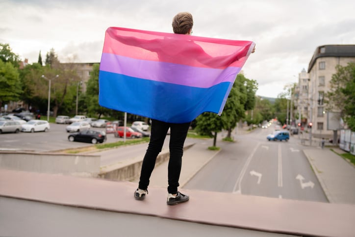 Man standing on an overpass holding the Bisexual flag, which is three bars of color on a rectangular field; the bar at the top is pink, the bar at the bottom is blue, and the center, where they overlap, is purple, to represent both hetero- and homosexual attraction