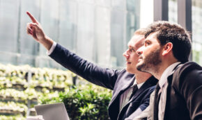 Image of two business-people in conversation: one is pointing at something through the window while the other looks up at it.