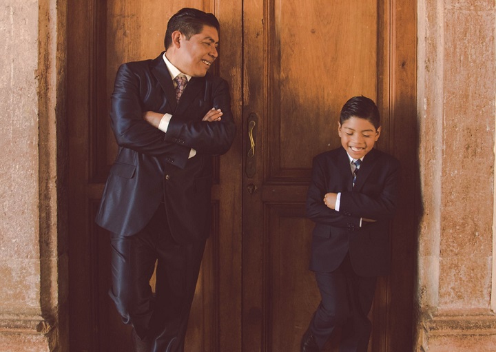 Photo of a middle aged father and pre-teen son, wearing matching tuxedos, making the same pose while their backs rest against a wooden door.