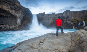 Image shows a natural waterfall just left of the photo's center. The fall spills into a wide, deep river. On the near riverbank, a man is standing, observing the waterfall. He is wearing a bright red jacket and blue jeans. The sky is blue and partially cloudy.