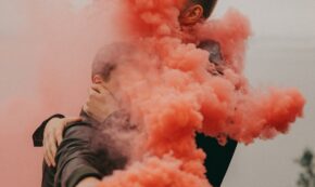 Two men, whose faces are obscured by a cloud of pink smoke, embrace in the street, celebrating.