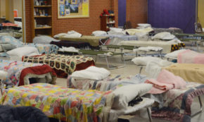 Image shows the inside of a temporary homeless shelter where several cots are made up with pillows and blankets, ready to receive the overnight houseless people who will sleep there for the night.