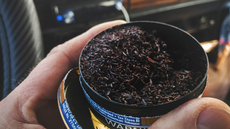 tin full of chewing tobacco, held in the hand of a man sitting behind the driver's wheel inside a car