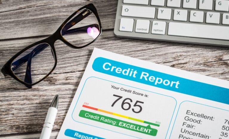 Image shows a credit report printed in color, with a score of 765. The report is on a desktop in front of a keyboard, and next to a pair of black-framed reading glasses.