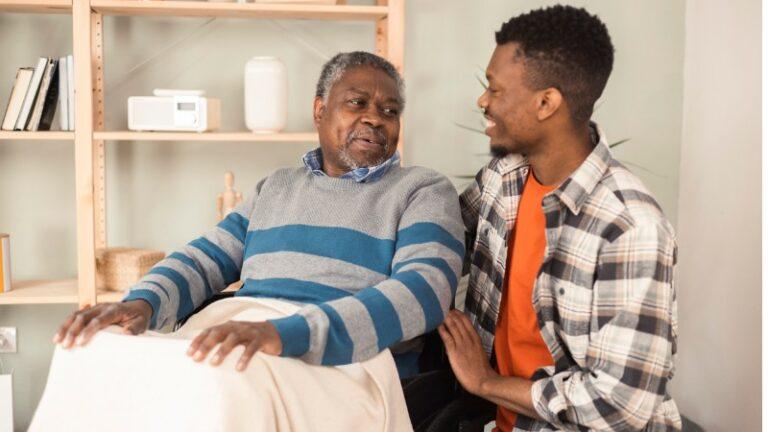 Photo description: Two African American men are shown. On the left is a senior sitting in a wheelchair. He is wearing a grey and blue wide-striped sweater over a button-down shirt, and he has a blanket over his lap. To the right in the photo, a younger man kneels at the older man's left. He has his right arm wrapped around the older man, and he is smiling. The younger man is wearing an orange t-shirt under an unbottoned brown and beige shirt. The two appear to have a familiar relationship such as father and son or uncle and nephew.