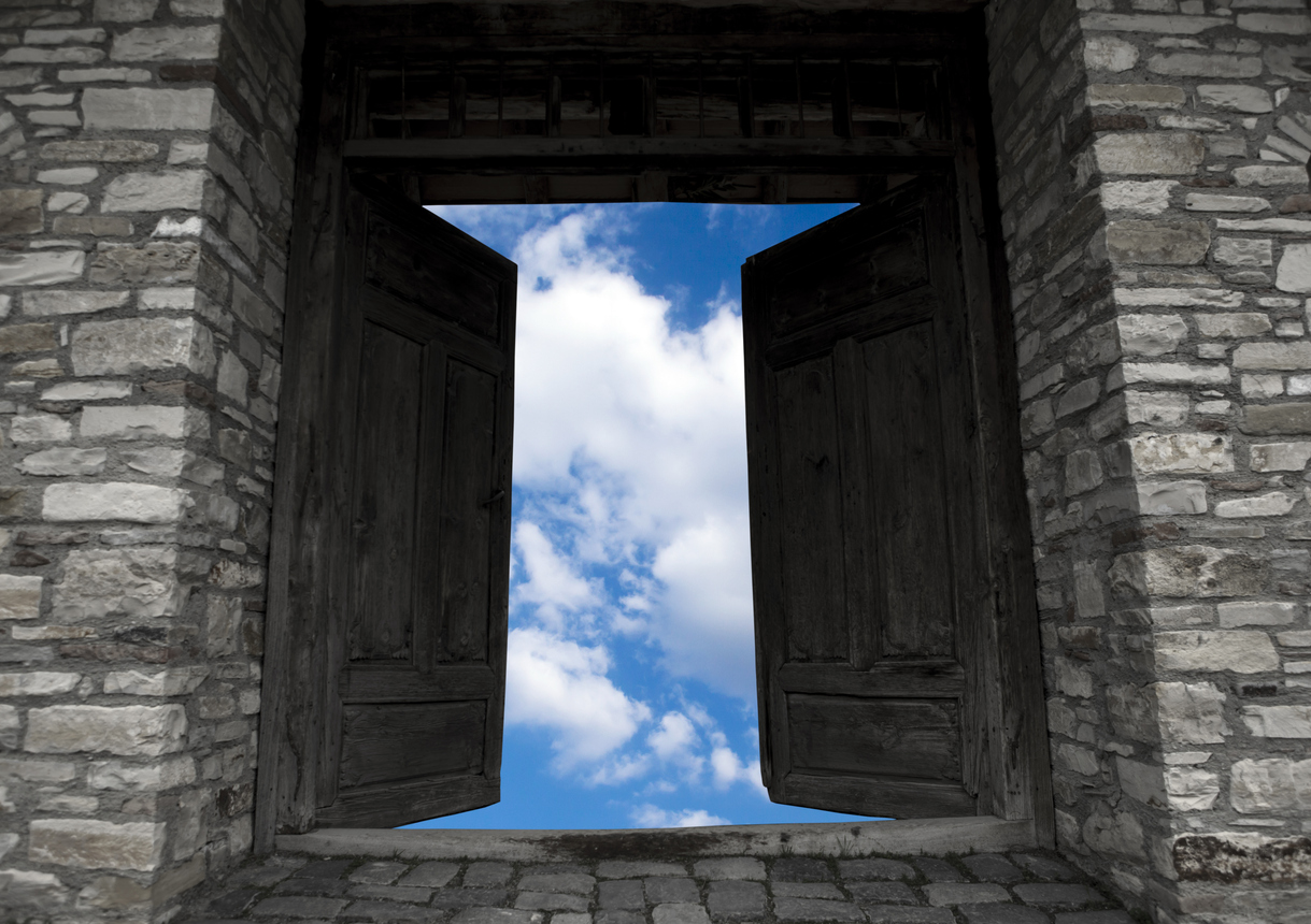 Photo shows two old dark wood castle doors framed by the stone building opening outward, revealing a bright blue sky with fluffy white clouds. There is stark contrast between the two elements.