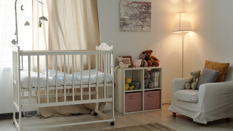 Interior of baby nursery with crib, storage cubie for toys, a floor lamp, and a comfortable chair with pillows. The overall color is white. The window is covered with white sheers, allowing the sun to fill the room.