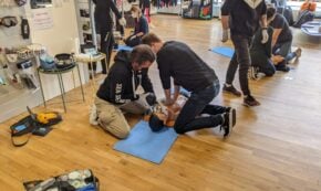 photo shows adults learning first aid and CPR. In the foreground, two people kneel at the sides of resusci anne cpr doll to simulate CPR on a blue rectangular matt over the wooden floor of the sports equipment store. The people are wearing casual clothing, mostly faded black and grey. One man wears beige khakis.
