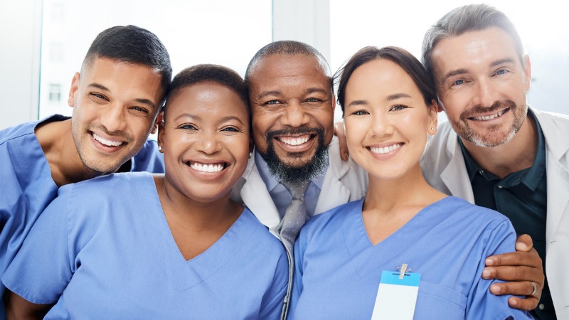 cheerful group of 5 diverse doctors standing with their arms around each other inside of a hospital during the day.