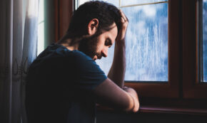 Photo shows a man with his head down in front of a window as he is leaning on the window sill at his elbows. It is raining outside and the clouded daylight is all that lights the man's face. His left hand is clenched lightly at his forehead and his right hand is holding his left elbow. He looks sorry or remorseful.