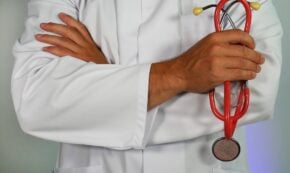 Photo shows a close-up of the chest only of a male medical doctor with light brown skin tone wearing a white coat (doctor's lab jacket), with his arms crossed, and his right hand holding a red stethoscope.