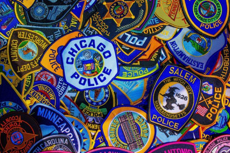 Photo shows several dozen police patches (such as those to be worn on a uniform shirt or jacket), each from a different municipality, with Chicago Police and Salem Police being prominently displayed. The photo is very colorful with the entire height and width being full of patches.