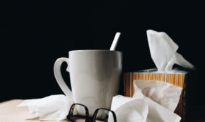 Image shows a white mug, black reading glasses, and a box of tissue all against a black background. The image relays the concept of someone being sick with a cold.