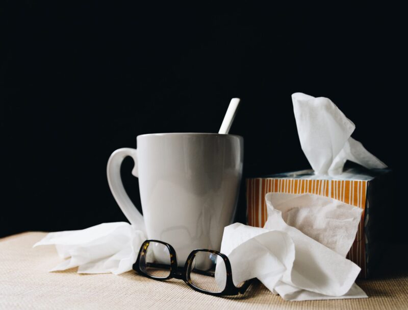 Image shows a white mug, black reading glasses, and a box of tissue all against a black background. The image relays the concept of someone being sick with a cold.