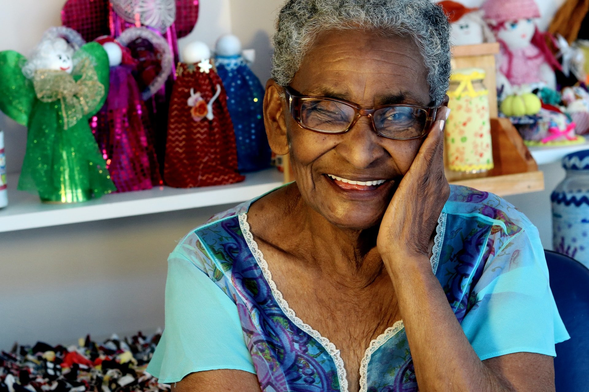 Elderly woman with medium brown skin and gray hair has a big smile with a full set of white teeth. She is wearing a 2-toned blue shirt and has her left hand on the side of her face, The background has colorful doll costumes and related crafts.