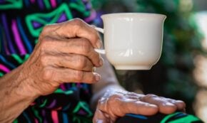 Image shows the hands of an elderly woman holding a white ceramic coffee cup. The woman is wearing a very colorful dress in a vibrant print. Her face is not shown in the image.