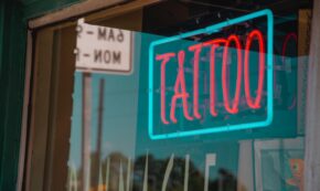 Image shows a neon sign in a shop window. The sign reads TATTOO in red capital letters framed by a teal rectangular border. Reflected in the shop window is part of a parking sign; we see only 6AM MON.