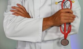 Image shows a close up of a male physician from below the neck to above the waste. The doctor is wearing a white clinic coat, has his arms folded across his chest, and is holding a red stethoscope.