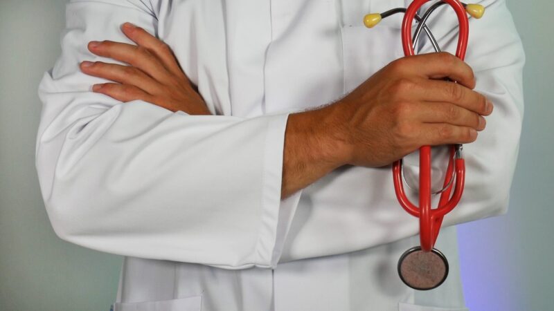 Image shows a close up of a male physician from below the neck to above the waste. The doctor is wearing a white clinic coat, has his arms folded across his chest, and is holding a red stethoscope.