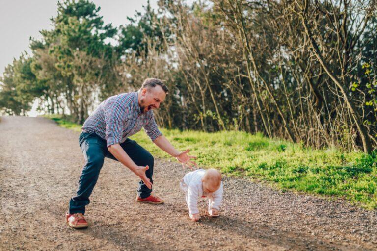 A father encourages his young toddler to take his first steps. The two are outdoors on a sunny day.