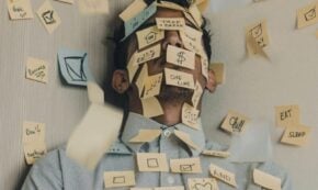 Image shows a young man up against the interior corner of a room with white walls. Covering the man's face and part of his blue dress shirt are small sticky notes, mostly yellow and some blue. Many notes are also stuck to the walls. The notes show various messages including "CHILL", "eat", "sleep", and the like, along with simple graphics such as that of a house, a heart, a cell phone, and "$$". Together these notes indicate stressors that are overwhelming the young man and pushing him up against the wall literally and figuratively.