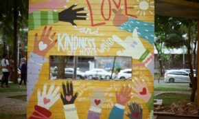 Image shows an outdoor divider wall painted with a yellow background on which are painted hands of children of different skin tones, each with a heart painted in the palm. The phrase "Love and Kindness are never wasted" is painted in the top half of the wall, with LOVE being in red and the rest in white paint.