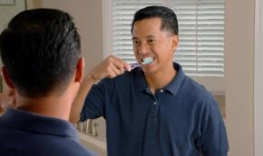 Image shows a light skinned man of maybe 35 years, likely of Asian descent, with short, straight, dark hair brushing his teeth while facing the bathroom mirror in a home. He is wearing a short-sleeved dark blue button-down shirt.