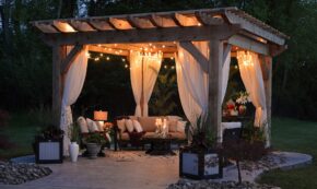 Image shows a nighttime summer backyard party setup just before the guests arrive. There is a canopy with white curtains tied to each corner post, comfortable seating, and soft lighting at the top edges of the canopy. The floor appears to be concrete with an outdoor area rug in a light gray color.