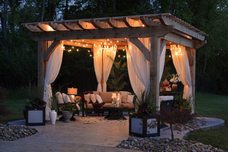 Image shows a nighttime summer backyard party setup just before the guests arrive. There is a canopy with white curtains tied to each corner post, comfortable seating, and soft lighting at the top edges of the canopy. The floor appears to be concrete with an outdoor area rug in a light gray color.