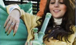 Image shows a mid-20s woman with light skin and long dark hair lounging, holding a silicone bong in her left hand. She is smiling in anticipation of enjoying the contents.