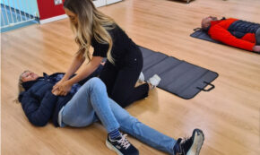Image shows CPR training in action. One person is practicing CPR on a human subject on a light-colored wood floor. Another human subject is in the background available for another CPR student.