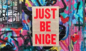 Image shows a sign JUST BE NICE in red letters on what seems to be a paper bag pasted to an outdoor wall of colorful graffiti.