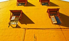 Image shows a close-up view of a three-story concrete home and 4 single windows, each with an orange awning. The house is painted a deep yellow. POV: We are standing so close to the building that we only see a tiny sliver of the blue sky on either side of the roof, but the shadow cast from the awning tells us it is a sunny day.