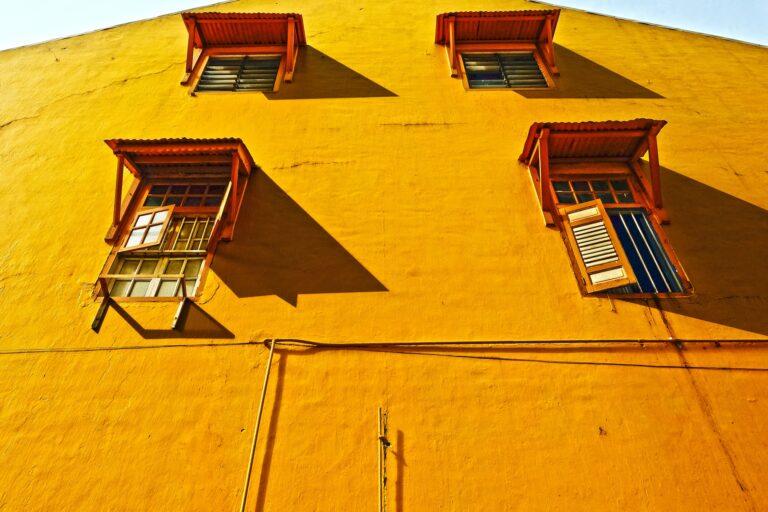 Image shows a close-up view of a three-story concrete home and 4 single windows, each with an orange awning. The house is painted a deep yellow. POV: We are standing so close to the building that we only see a tiny sliver of the blue sky on either side of the roof, but the shadow cast from the awning tells us it is a sunny day.