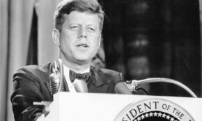 The black and white image shows US President John F. Kennedy, Jr. (Nov. 18, 1963) at a podium with the official presidential emblem.