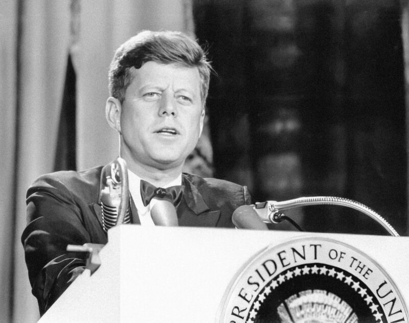 The black and white image shows US President John F. Kennedy, Jr. (Nov. 18, 1963) at a podium with the official presidential emblem.