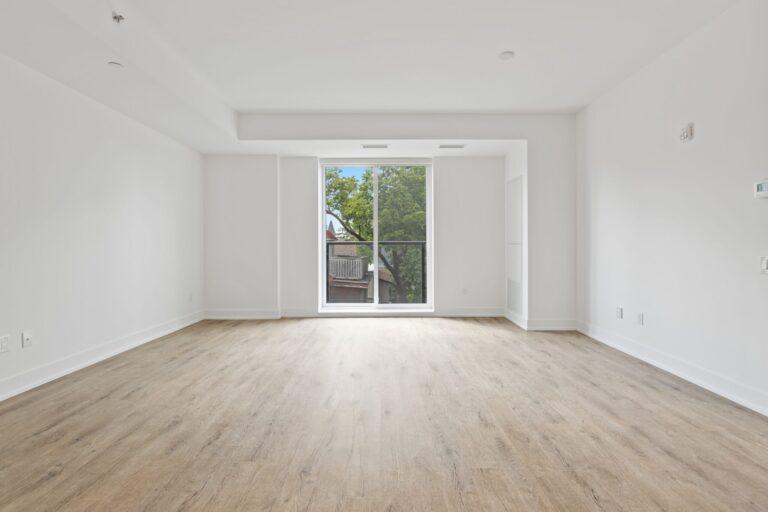 Image shows the inside view of an apartment's empty living room with a sliding glass door to a patio with live green plants. The clean-looking room has white walls and ceiling, and a light ash wood-look floor.