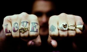 Image shows the knuckles of a light-skinned person showing their rings and tattoos. On the right hand is the word LOVE with a gold skull ring on the V finger. On the left hand are a spade tattoo and three rings.