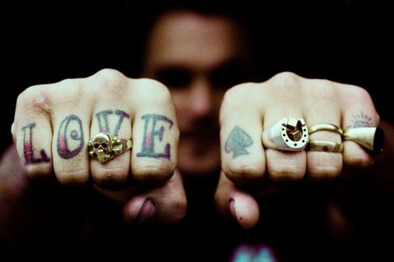 Image shows the knuckles of a light-skinned person showing their rings and tattoos. On the right hand is the word LOVE with a gold skull ring on the V finger. On the left hand are a spade tattoo and three rings.