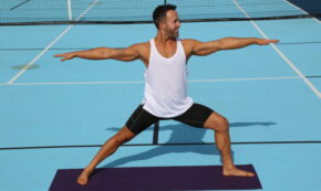 Photo shows fitness expert Julian Brass doing a yoga pose. He is wearing a white tank top and black shorts. The background is blue.