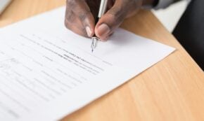 Image shows a close-up of the right hand of a person signing a contract with a nice pen. The document is on a light-colored wooden table.